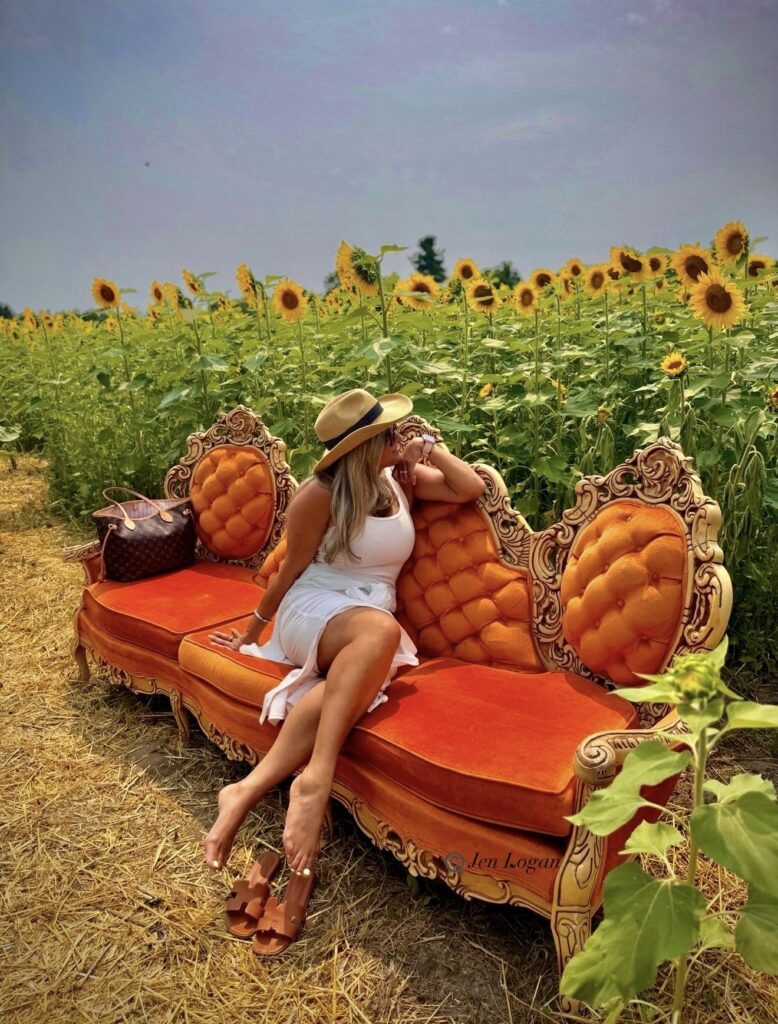 Woman poses on a prop antique orange couch with ornate wood carving trim in the middle of a sunflower field at B'More Sunflower Farm in Whitby
